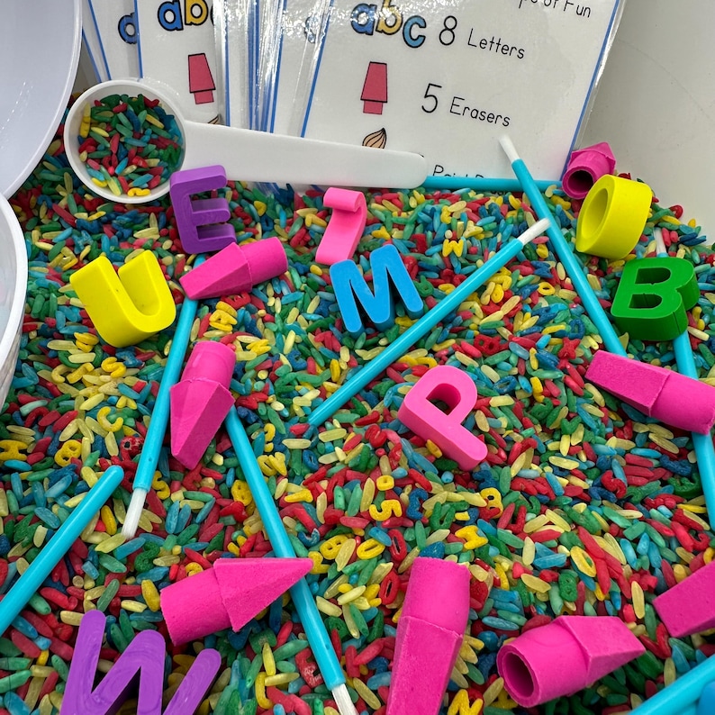 May include: A sensory bin filled with colorful rice, letters, and foam erasers. The bin includes blue, pink, yellow, green, and purple letters, as well as blue and pink erasers. The bin also includes a white scoop, blue sticks, and a white paintbrush.