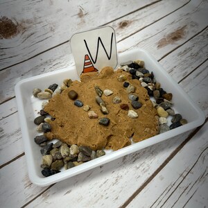 May include: A white plastic tray containing brown playdough and an assortment of small, coloured pebbles. A small card with the letter "W" and a traffic cone illustration is placed in the playdough. The background is a light wood grain.