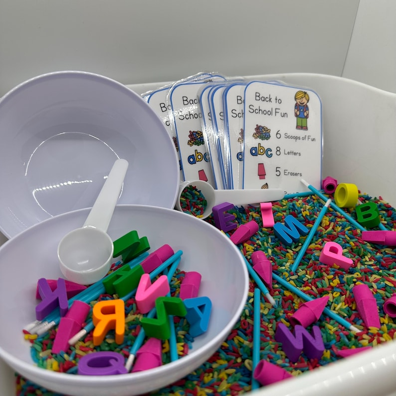 May include: A white bowl filled with colorful rice, alphabet letter blocks, and blue and pink sticks. Two smaller white bowls are on top of the larger bowl. A white scoop is next to the bowls. A stack of cards with the text "Back to School Fun" is in the background.