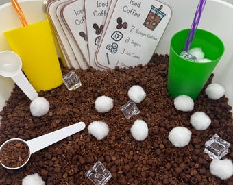 May include: A sensory bin with a coffee theme. It includes a yellow cup with an orange straw, a green cup with a purple straw, white scoops, and cards that say "Iced Coffee" with a recipe. The bin is filled with brown textured material, white cotton balls, and clear plastic ice cubes.