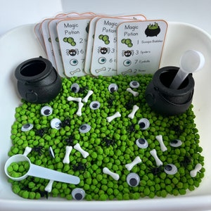 May include: A white bowl filled with green sensory beans, black spiders, white bones, and googly eyes. There are four cards with the words "Magic Potion" and numbers on them. There are two black cauldrons with white scoops.