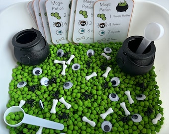 May include: A white bowl filled with green sensory beans, black spiders, white bones, and googly eyes. There are four cards with the words "Magic Potion" and numbers on them. There are two black cauldrons with white scoops.