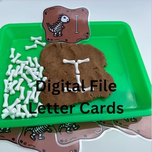 May include: A green tray holds a brown playdough shape with the letter "I" formed from white bone-shaped pieces. A card with a dinosaur skeleton and the letter "I" sits above the playdough. The text "Digital File Letter Cards" is below.