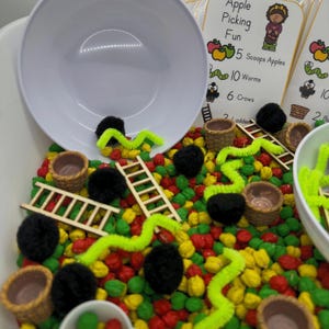 May include: A sensory play setup with a white bowl, small wooden ladders, and miniature woven baskets. The scene includes colorful beads, black pom-poms, and bright green pipe cleaners. Cards with the text "Apple Picking Fun" are also visible.