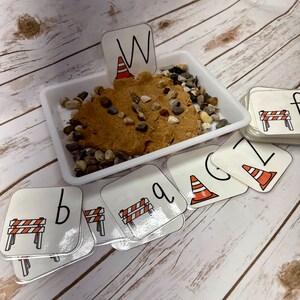 May include: A white tray containing brown playdough, small stones, and letter cards. The letter cards feature a construction theme with images of traffic cones and barriers. The letter "W" is placed in the playdough.