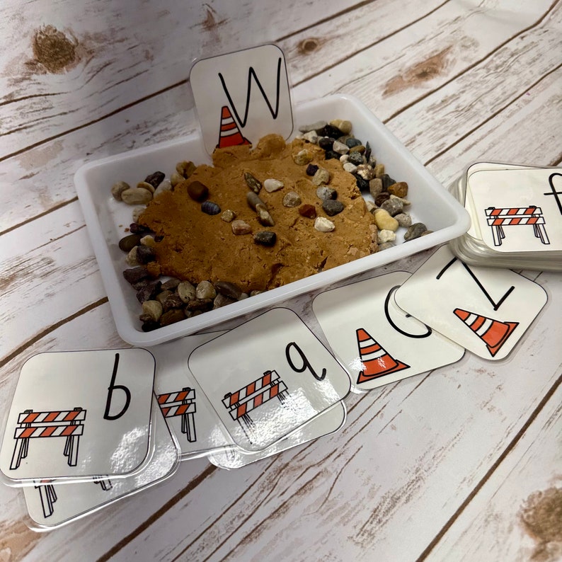 May include: A white tray containing brown playdough, small stones, and letter cards. The letter cards feature a construction theme with images of traffic cones and barriers. The letter "W" is placed in the playdough.