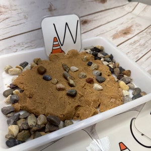 May include: A sensory play setup in a white tray. It features a brown, textured substance resembling sand, adorned with various small pebbles. A card with the letter "W" and an orange traffic cone graphic is placed in the sand.