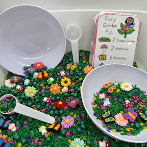 May include: A white plastic container filled with green rice, colorful sprinkles, and small plastic figurines of butterflies, mushrooms, and flowers. A white card with the text "Fairy Garden Fun" and a list of items needed for the activity is visible in the background.