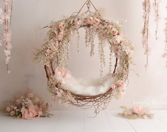 May include: A white, fluffy, circular nest made of twigs and vines, decorated with pink flowers and greenery, hangs from the ceiling. The nest is surrounded by pink flowers and greenery, and the background is a light pink wall.