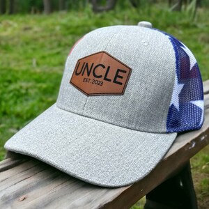 May include: A gray and white baseball cap with a brown leather patch that says "UNCLE EST. 2023". The back of the cap has a blue and white mesh panel with a star pattern.