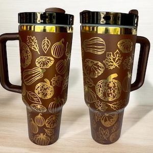 May include: Two brown travel mugs with gold accents and a autumn-themed design featuring pumpkins, leaves, and gourds. The mugs have a handle and a lid.