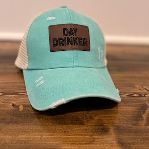 May include: A turquoise blue and white distressed trucker hat with a brown leather patch that says "DAY DRINKER" in black lettering.