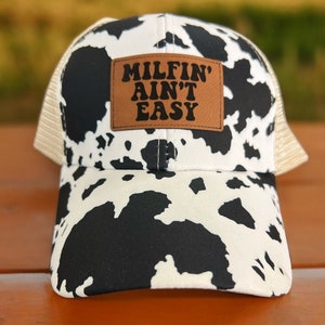 Op de afbeelding: Een zwarte en witte koeienprint trucker cap met een bruin leren patch waarop "MILFIN' AINT' EASY" staat.