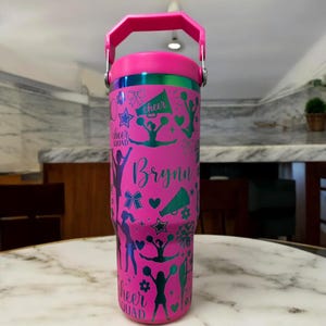 Pode incluir: Um copo rosa brilhante com alça e tampa, apresentando um padrão iridescente verde metálico de silhuetas de líderes de torcida, megafones, laços, estrelas e corações. O nome Brynn está impresso em letra cursiva, com o texto Cheer Squad.