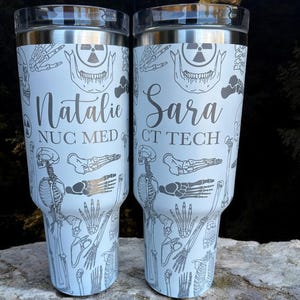 May include: Two white stainless steel tumblers with a skeletal design and personalized names. The tumbler on the left says "Natalie NUC MED" and the tumbler on the right says "Sara CT TECH".