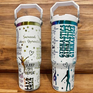 Può includere: Due bicchieri isolanti bianchi con manici, decorati con grafica e testo a tema ginnastica. Un bicchiere presenta il nome "Savannah" e frasi come "Eat Sleep Gym Repeat". L'altro ha "Gymnastics" ripetuto verticalmente.