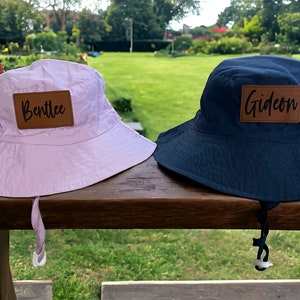 May include: Two personalised bucket hats. One is pale pink with the name "Bentlee" on a brown patch. The other is navy blue with the name "Gideon" on a brown patch. Both hats have a chin strap and are on a wooden surface.