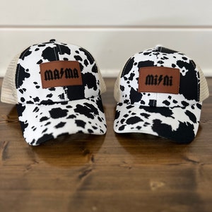 Può includere: Due cappellini da camionista con stampa a mucca bianca e nera con toppe in pelle marrone. La toppa sul cappello a sinistra dice "mama" e la toppa sul cappello a destra dice "mini".