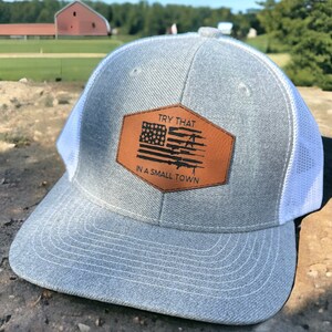May include: Gray and white trucker hat with a brown leather patch featuring an American flag and the text "TRY THAT IN A SMALL TOWN".