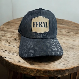 Op de afbeelding: Zwarte truckerpet met een luipaardprint op de klep en een mesh achterkant. De pet heeft een bruinbeige patch met het woord "FERAL" in vette, zwarte letters.