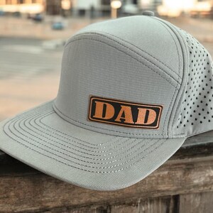 May include: A grey and black baseball cap with a brown leather patch that says "DAD" in orange letters.