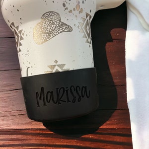 May include: White travel mug with a gold and black geometric pattern, a gold cowboy hat with leopard print, and a black silicone bottom with the name "Marissa" printed on it.