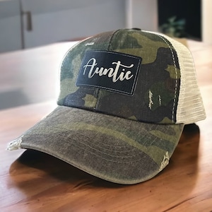 Può includere: Un cappello da camionista in rete bianca e mimetica con una toppa nera che dice "Auntie" in corsivo bianco.