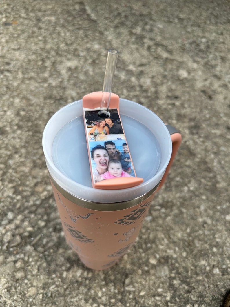 May include: A pink travel mug with a geometric pattern and a clear lid. The lid has a slot for a straw and a photo strip with pictures of a family.