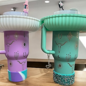 Könnte beinhalten: Zwei farbenfrohe Reisebecher mit Deckeln und dekorativen Designs. Die Becher sind lila und grün mit silbernen Akzenten. Die Becher haben ein Western-Thema mit Kaktus, Sternen und einem Totenkopf-Design.