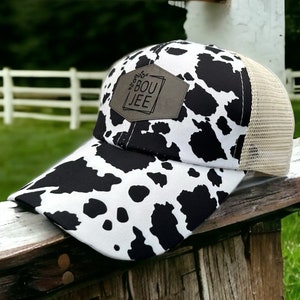 Può includere: Un cappello da camionista con stampa di mucca bianco e nero con una toppa in pelle marrone che dice "BOU JEE".