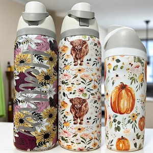 May include: Three insulated water bottles with floral, highland cow, and pumpkin designs. The bottles have white and gray lids. One bottle has the name "Madison" in silver script. The other two bottles have a floral pattern with a highland cow and a pumpkin.