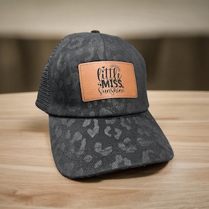 May include: A black and grey leopard print trucker hat with a brown leather patch that says "little miss sunshine".
