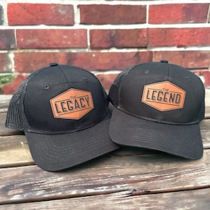 May include: Two black trucker hats with brown leather patches. The patch on the left hat says "The Legacy" and the patch on the right hat says "The Legend".