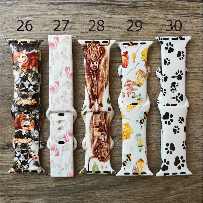 May include: Five different Apple Watch bands with various colorful patterns. The bands are numbered 26 through 30. The patterns include cats, flamingos, a Highland cow, bees and honeycombs, and paw prints.