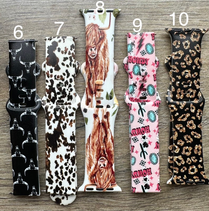 May include: Five different Apple Watch bands with various patterns. The bands are numbered 6 through 10. Band 6 is black with white skulls, band 7 is white with brown and black spots, band 8 is white with a brown Highland Cow, band 9 is pink with turquoise and white lightning bolts and the word "Howdy" repeated, and band 10 is black with gold glitter and leopard spots.