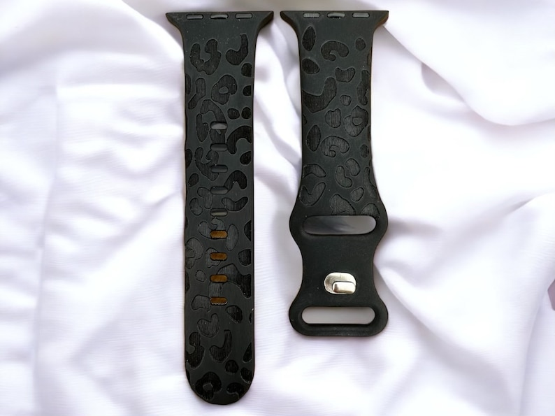 Pu&ograve; includere: Un paio di cinturini per Apple Watch in silicone nero con un design a stampa leopardata. I cinturini sono mostrati su una superficie bianca.