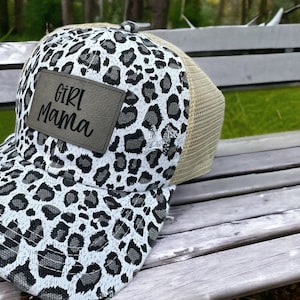 Può includere: Un cappello da camionista con stampa leopardata bianco e nero con una toppa in pelle marrone che dice "Girl Mama".