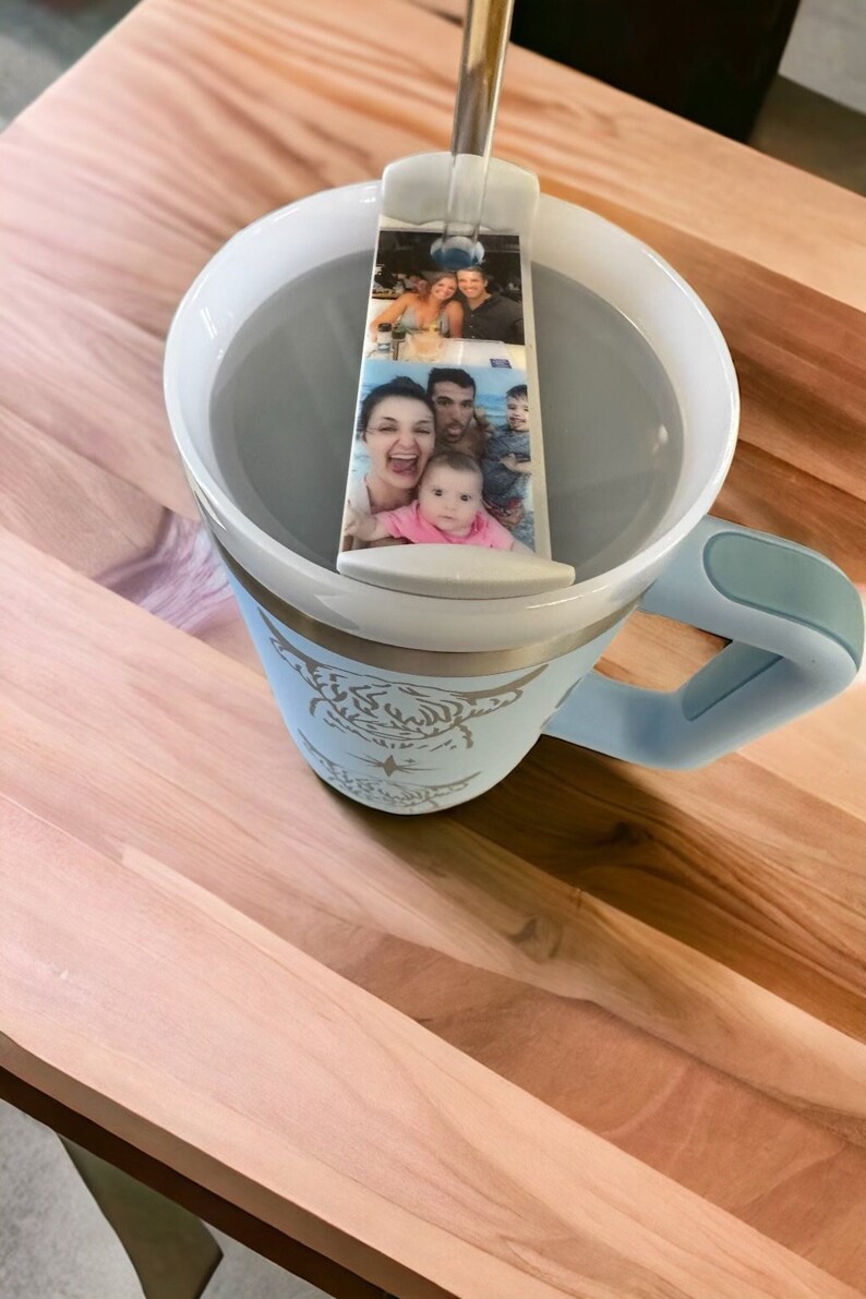 May include: A light blue travel mug with a white lid and a silver band. The mug has a design of a cow on the front. A photo strip of a family is attached to the lid.