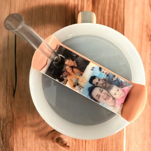 May include: A white ceramic mug with a gray lid and a clear plastic straw. The lid has a pink silicone band with a photo of a family printed on it.