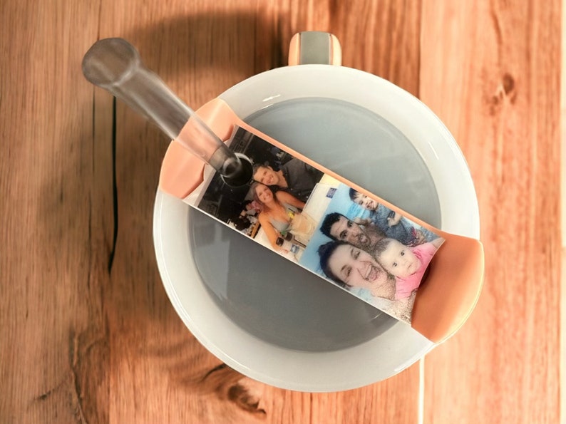 May include: A white ceramic mug with a gray lid and a clear plastic straw. The lid has a pink silicone band with a photo of a family printed on it.