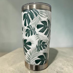 Op de afbeelding: Een cilindervormige roestvrijstalen beker met een witte achtergrond en een herhalend patroon van donkergroene monsterabladeren en lichtgrijze palmbladeren. De beker heeft een metallic zilveren rand aan de bovenkant en een bijpassende zilveren voet.