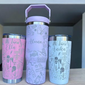 Könnte beinhalten: Drei Edelstahlbecher mit medizinischem Thema. Die Becher sind mit den Namen "Aleena", "Ella" und "Anika" personalisiert. Die Becher sind in den Farben Rosa, Lila und Blau.