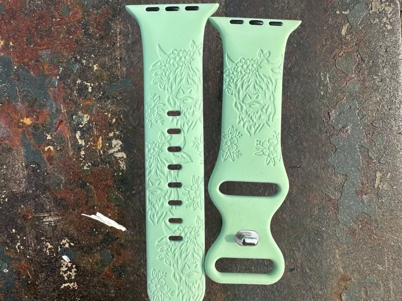 May include: A pair of light green Apple Watch bands. Each band features a detailed floral and Highland cow design. The bands are made of a flexible material and have a silver clasp. The bands are designed to fit a standard Apple Watch.
