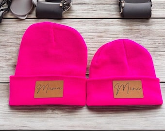 May include: Two pink knit beanies with brown leather patches. The larger beanie has the word "Mama" on the patch and the smaller beanie has the word "Mini" on the patch.