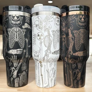 Könnte beinhalten: Drei Edelstahlbecher mit einem Skelett- und Strahlungssymbol-Design. Die Becher sind schwarz, weiß und kupferfarben.