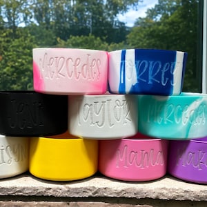 以下が含まれることがあります： 「メルセデス」「ジェニ」「テイラー」「メルセデス」「トーリ」「マディソン」「ママ」「マケイリン」など、さまざまな名前が刻まれた、カラフルなシリコン製のカップホルダー9個が積み重ねられています。