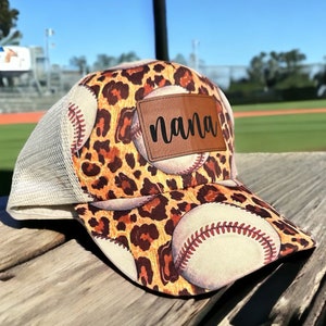 Può includere: Un cappello da baseball beige con stampa leopardata e disegni di baseball. Il cappello ha una toppa di pelle marrone con la parola "nana" su di essa.