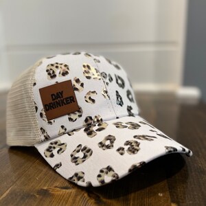 Può includere: Cappellino da baseball bianco con motivo leopardato e retro in rete. Il cappellino presenta una toppa in pelle marrone con la scritta "DAY DRINKER". La visiera e i pannelli anteriori mostrano un motivo ripetuto di macchie marroni e nere.