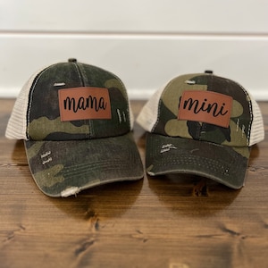 Può includere: Due cappellini da camionista mimetici con toppe in pelle marrone. La toppa sul cappello a sinistra dice "mama" e la toppa sul cappello a destra dice "mini".