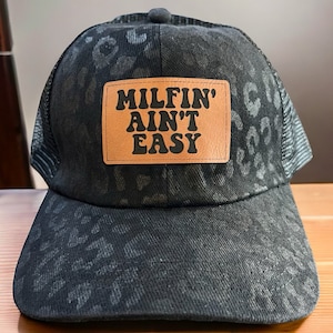 May include: Black trucker hat with a leopard print pattern and a brown patch that says "MILFIN' AINT' EASY" in black letters.
